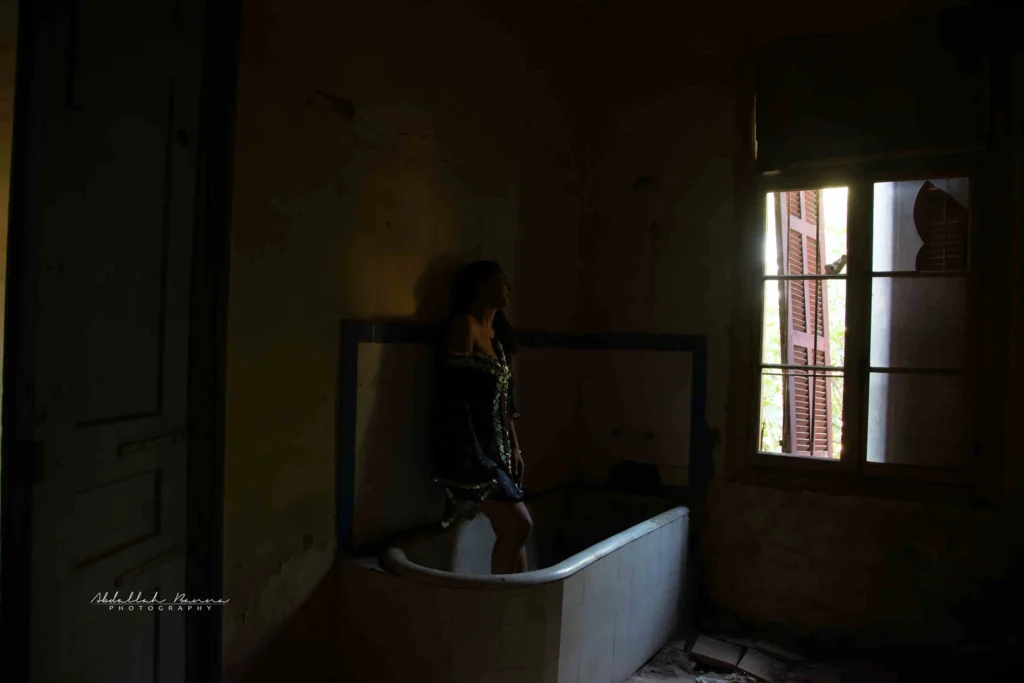 Woman standing in an old bathtub in a dimly-lit abandoned bathroom, looking out the window.