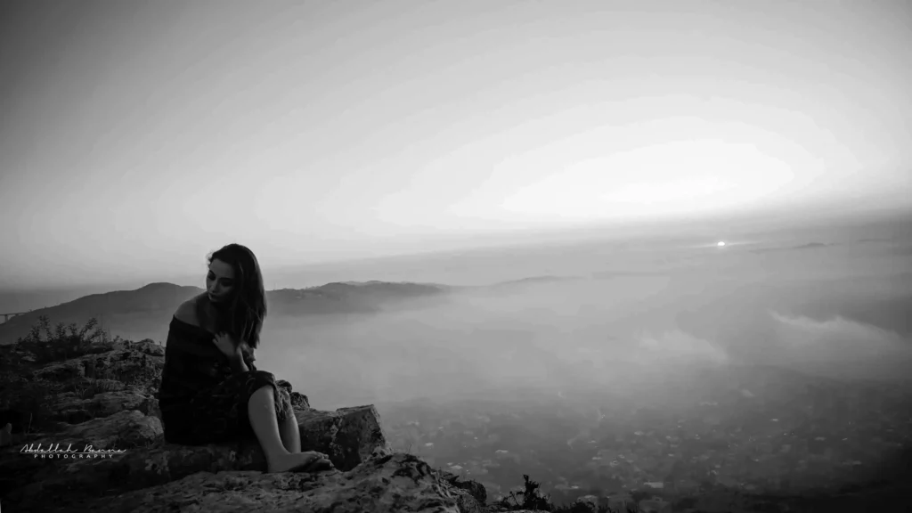 Black and white photo of woman sitting on cliff edge with misty landscape.