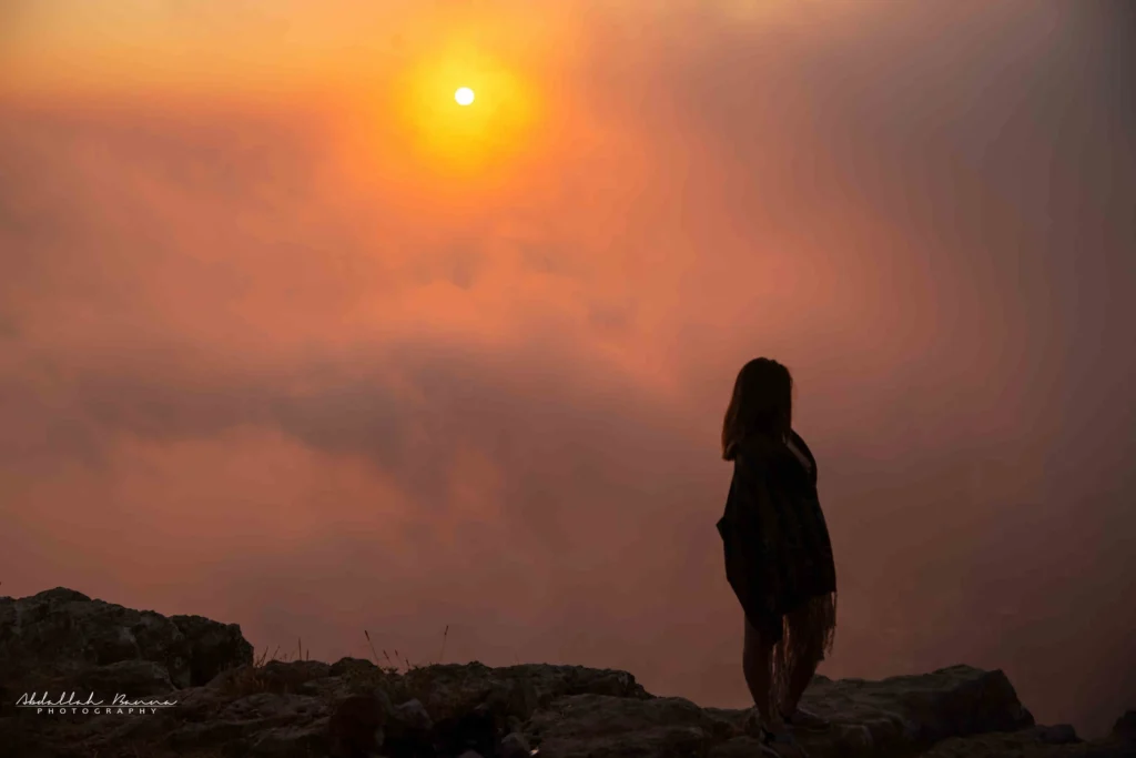 Silhouette of woman standing on rocky cliff during sunset with thick clouds.