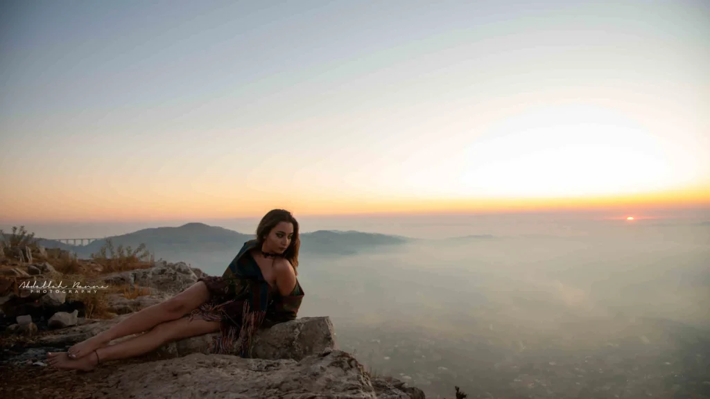 Woman sitting on rocks with a sunset and misty mountains in the background.