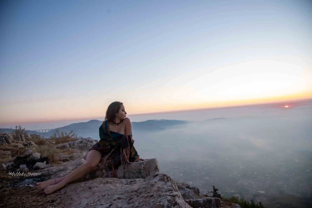 Woman wrapped in shawl sitting on rocky ledge watching the sunset.