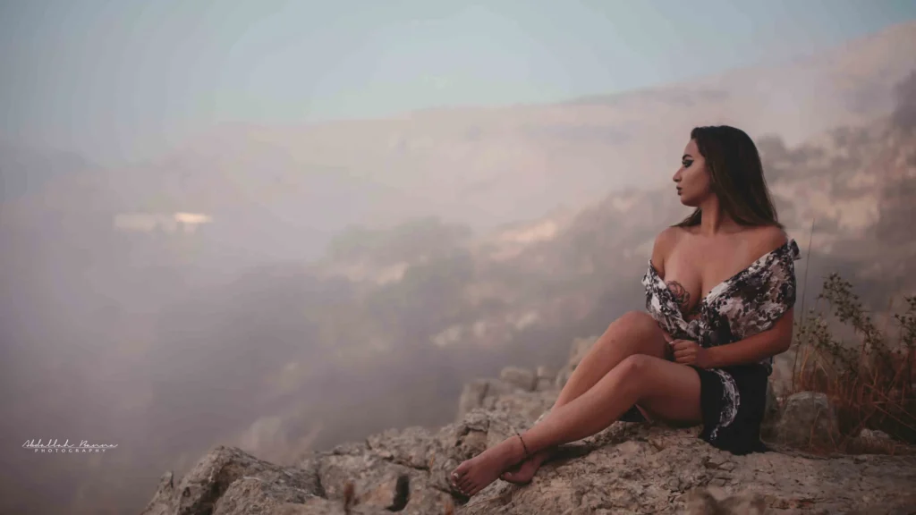 Woman sitting by an arched window looking at green foliage outside in a dimly-lit room.