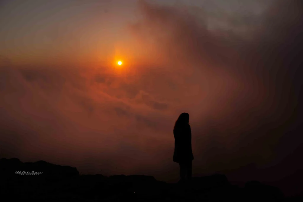 Woman standing on rocky outcrop looking at bright setting sun in misty orange sky.