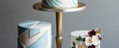 Instead of a traditional stacked cake, this design features three separate tiers displayed on individual gold stands. Each tier has a different design: one with white fondant and geometric shapes, another with marbled fondant in soft blues, and the third with a floral motif. The staggered heights of the tiers create a modern, asymmetrical presentation.