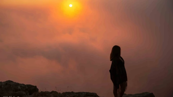 Silhouette of woman standing on rocky cliff during sunset with thick clouds.