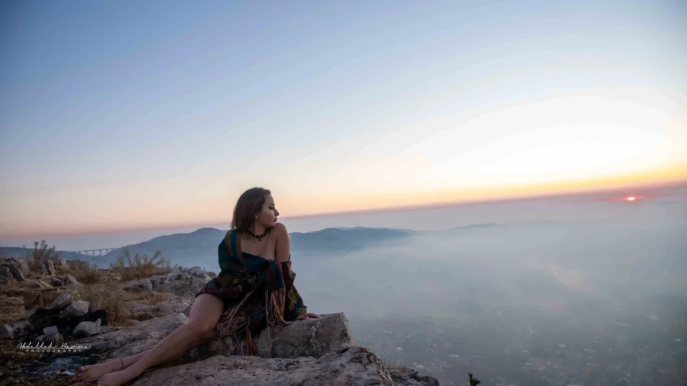 Woman wrapped in shawl sitting on rocky ledge watching the sunset.