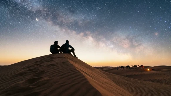 لقطة سينمائية لصديقين يجلسان فوق كثبان رملية تحت سماء مرصعة بالنجوم تعبيراً عن رفيق الدرب